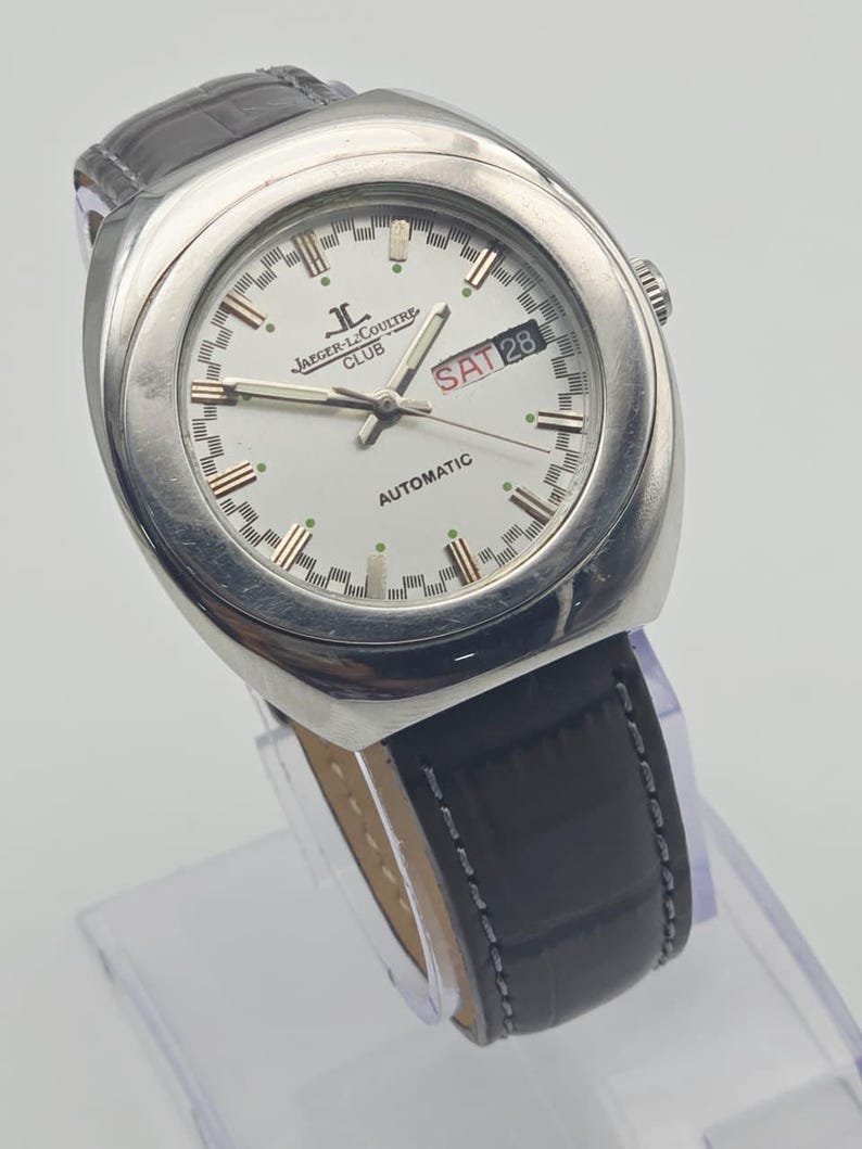 Puede incluir: Un reloj autom&aacute;tico Jaeger-LeCoultre Club plateado con esfera blanca, correa de cuero negro y la fecha mostrada como S&Aacute;B 28. El reloj tiene agujas e indicadores de hora plateados, y la correa es texturizada.