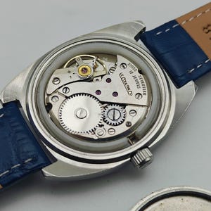 Puede incluir: Un reloj de pulsera vintage sin la parte trasera, que revela el intrincado movimiento mec&aacute;nico. El reloj tiene una caja plateada, una correa de cuero azul y el texto "SWISS 17 JEWELS LE COULTRE CO."