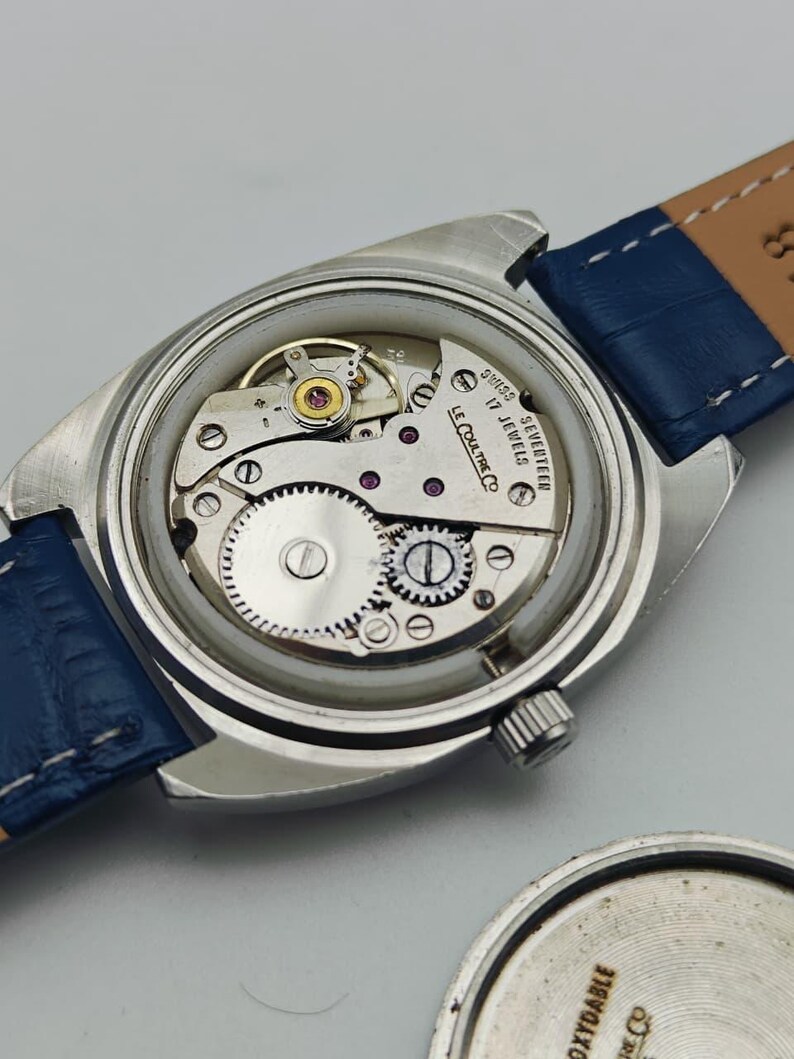 Puede incluir: Un reloj de pulsera vintage sin la parte trasera, que revela el intrincado movimiento mec&aacute;nico. El reloj tiene una caja plateada, una correa de cuero azul y el texto "SWISS 17 JEWELS LE COULTRE CO."
