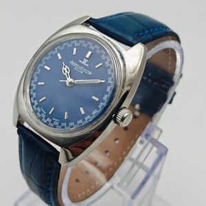 Puede incluir: Un reloj Jaeger-LeCoultre Club vintage con esfera azul y caja plateada. El reloj tiene una correa de cuero azul. La esfera muestra el logotipo de Jaeger-LeCoultre y la palabra "CLUB".