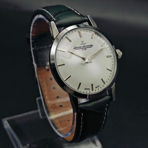 Pu&ograve; includere: Un orologio Jaeger-LeCoultre Club argentato con quadrante chiaro e lancette argentate. L'orologio ha un cinturino in pelle verde scuro ed &egrave; contrassegnato "Swiss Made". L'orologio &egrave; esposto su un supporto trasparente.
