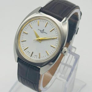 May include: A silver Ulysse Nardin wristwatch with a white face and gold hands and markers. The watch has a dark brown leather strap. The watch is displayed on a clear stand.