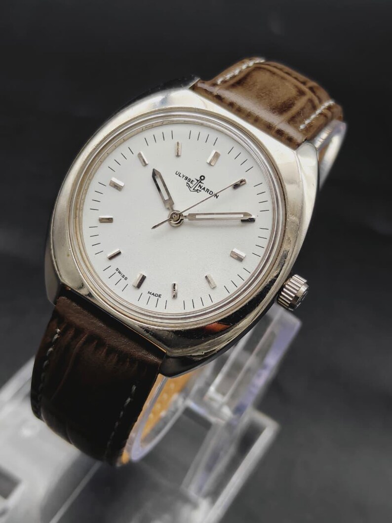 May include: A silver-tone wristwatch with a white dial, silver baton indices, and silver hands. The watch features the text Ulysse Nardin and an anchor logo below the twelve o'clock position, with Swiss Made at the bottom. It has a brown leather strap.