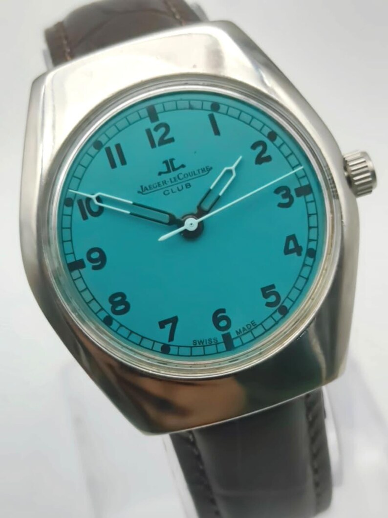 Op de afbeelding: Een zilverkleurig Jaeger-LeCoultre Club horloge met een turquoise wijzerplaat en zwarte cijfers. Het heeft een bruine leren band en de tekst "SWISS MADE".