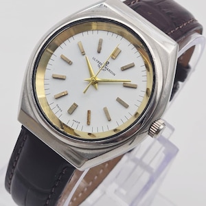 Pu&ograve; includere: Un orologio Ulysse Nardin argentato con quadrante bianco, dettagli dorati e cinturino in pelle marrone. L'orologio ha una cassa rettangolare e un design classico. Il quadrante mostra il nome del marchio e la scritta "SWISS MADE".