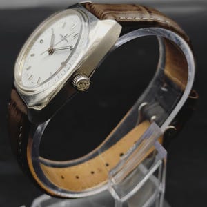 May include: A wristwatch with a silver-toned metal case and a white dial featuring silver hour markers and hands. The watch has a brown leather strap with a textured finish and a crown on the side embossed with the letters JN.