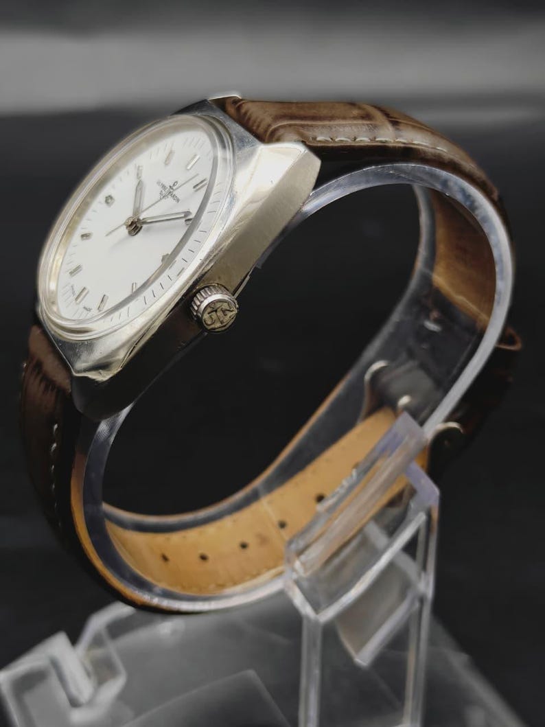 May include: A wristwatch with a silver-toned metal case and a white dial featuring silver hour markers and hands. The watch has a brown leather strap with a textured finish and a crown on the side embossed with the letters JN.