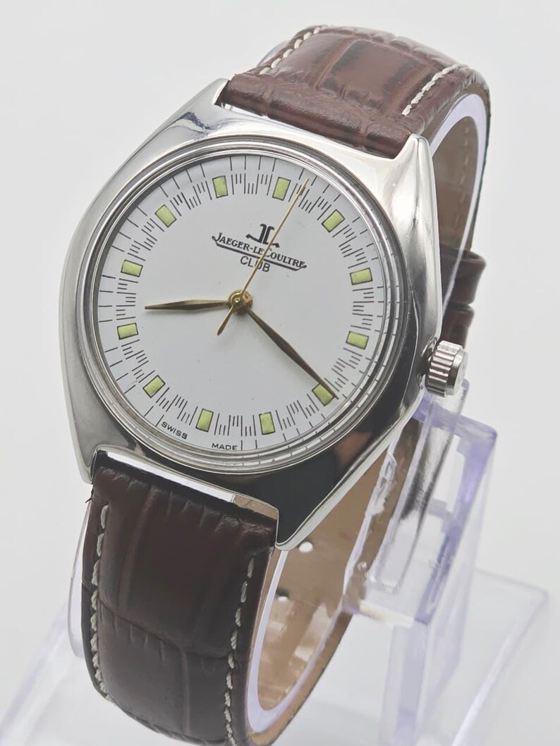 May include: A silver-toned Jaeger-LeCoultre Club watch with a white face, featuring the brand's logo and the words "SWISS MADE." The watch has a brown leather band with white stitching and gold-toned hands.