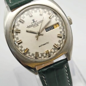 Puede incluir: Un reloj autom&aacute;tico Jaeger-LeCoultre Club plateado con esfera blanca y correa de cuero verde. La esfera del reloj muestra el d&iacute;a y la fecha. El reloj tiene una esfera redonda con agujas y marcadores plateados.