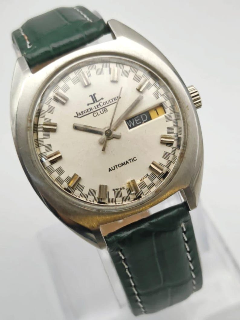 Puede incluir: Un reloj autom&aacute;tico Jaeger-LeCoultre Club plateado con esfera blanca y correa de cuero verde. La esfera del reloj muestra el d&iacute;a y la fecha. El reloj tiene una esfera redonda con agujas y marcadores plateados.