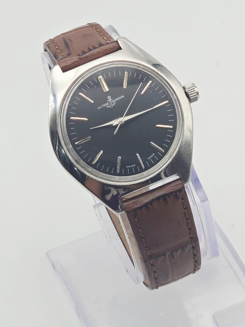 May include: A silver-toned wristwatch with a black face and brown leather band. The watch face features silver hour markers and hands. The band is a dark brown, textured leather. The watch is displayed on a clear stand.