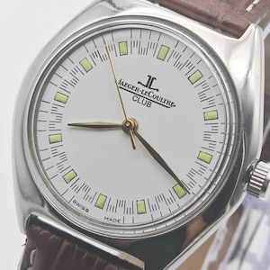 May include: A silver Jaeger-LeCoultre Club watch with a white face, gold hands, and a brown leather strap. The watch face features the brand name and the words "SWISS MADE".