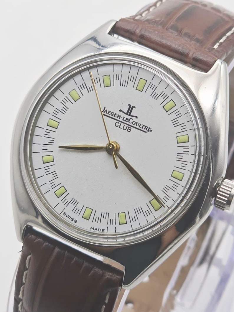 May include: A silver Jaeger-LeCoultre Club watch with a white face, gold hands, and a brown leather strap. The watch face features the brand name and the words "SWISS MADE".