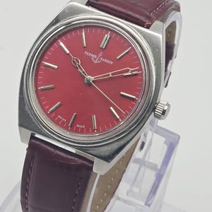May include: A vintage Ulysse Nardin watch with a vibrant red dial and silver hands. The watch features a stainless steel case and a burgundy leather strap. The watch is displayed on a clear stand.