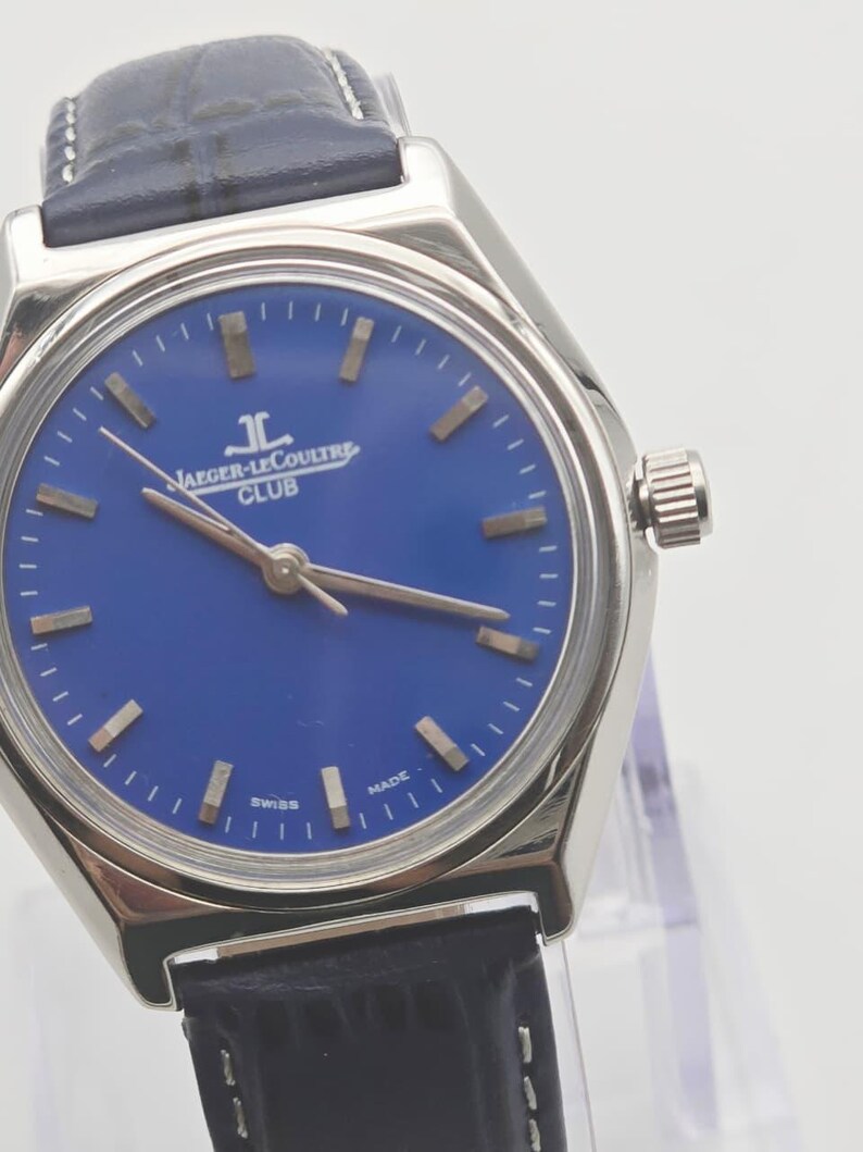 May include: A stainless steel Jaeger-LeCoultre Club watch with a blue dial and a black leather strap. The watch face features gold-colored hour markers and hands. The text "JAEGER-LECOULTRE CLUB" is printed on the dial. The watch is marked "SWISS MADE".