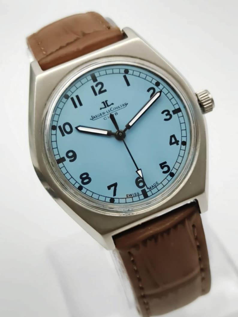 K&ouml;nnte beinhalten: Eine silberfarbene Armbanduhr mit hellblauem Zifferblatt und braunem Lederarmband. Das Zifferblatt zeigt schwarze Ziffern und Zeiger, mit der Marke "JAEGER-LECOULTRE" und "SWISS MADE". Die Uhr hat ein rechteckiges Geh&auml;use.