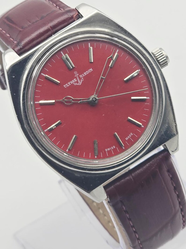 May include: A vintage Ulysse Nardin wristwatch with a red dial and silver-tone case. The watch features a burgundy leather strap and the brand's logo. The dial has silver markers and hands. The watch is marked "SWISS MADE".
