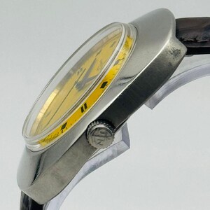 May include: A vintage wristwatch with a silver case and a yellow dial. The watch has black hour markers and hands, and a brown leather strap. The watch is angled, showing its side profile.