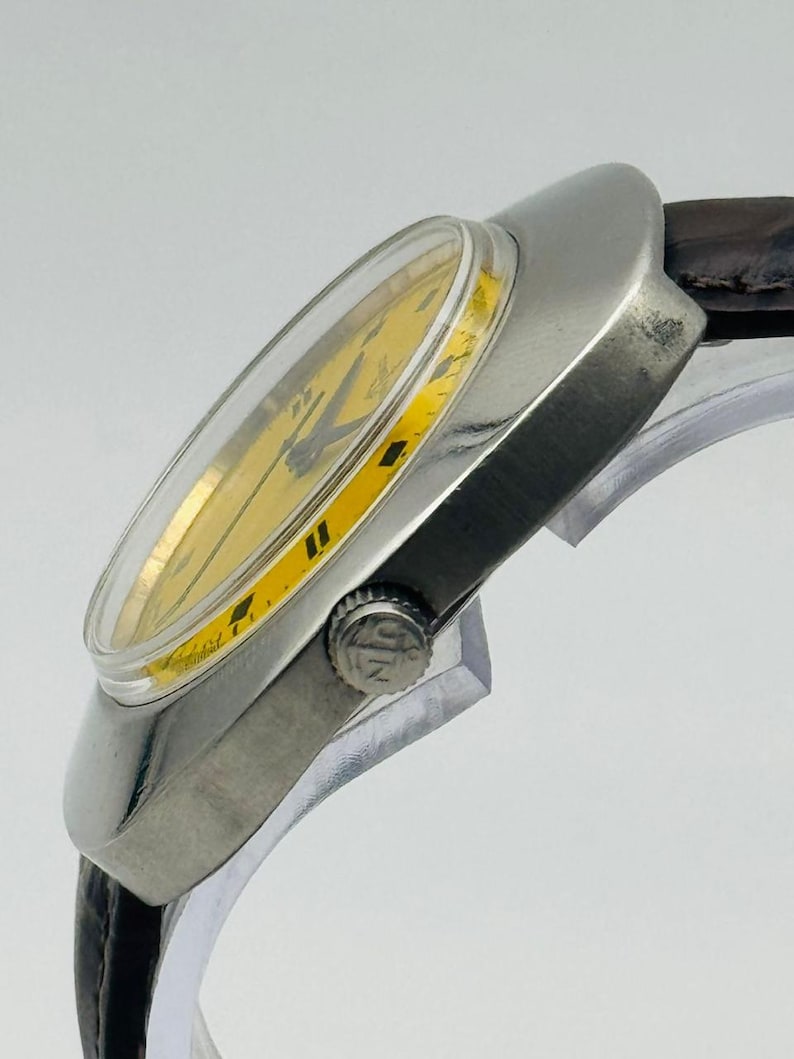 May include: A vintage wristwatch with a silver case and a yellow dial. The watch has black hour markers and hands, and a brown leather strap. The watch is angled, showing its side profile.