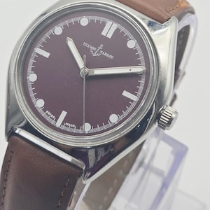 May include: A silver-toned wristwatch with a burgundy face and brown leather band. The watch face features white hour markers and hands, with the brand name "Ulysse Nardin" visible. The watch is displayed on a clear stand.