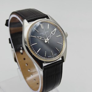 May include: A vintage Ulysse Nardin wristwatch with a dark blue face and silver hands. The watch has a silver case and a black leather band. The watch is displayed on a clear stand. The watch face has the text "Ulysse Nardin" and "Swiss Made".