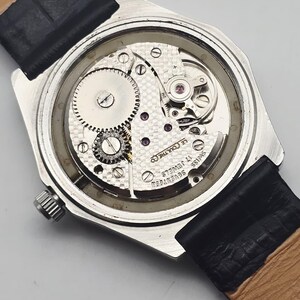 May include: A close-up view of the back of a vintage wristwatch, showcasing the intricate mechanical movement. The watch has a silver-toned case, a black leather strap, and the words "Swiss Seventeen 17 Jewels" are visible.