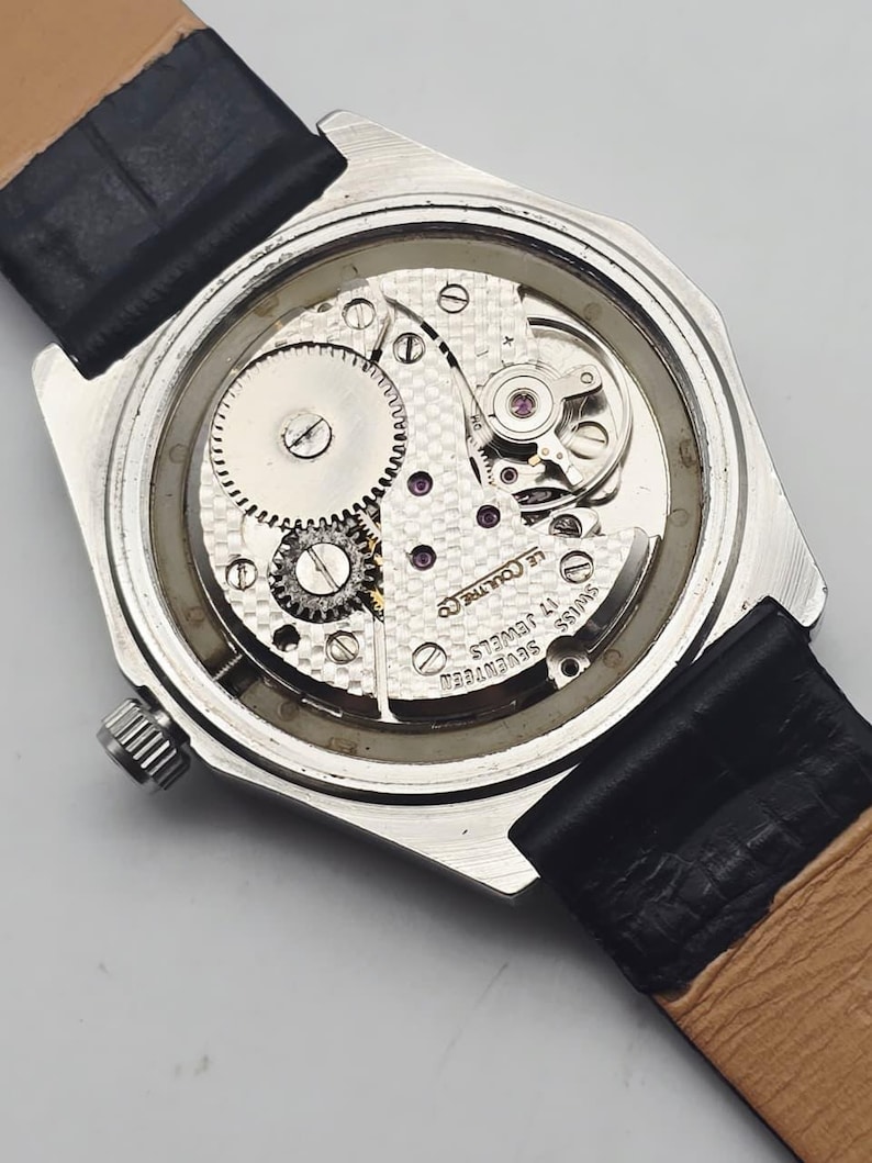 May include: A close-up view of the back of a vintage wristwatch, showcasing the intricate mechanical movement. The watch has a silver-toned case, a black leather strap, and the words "Swiss Seventeen 17 Jewels" are visible.