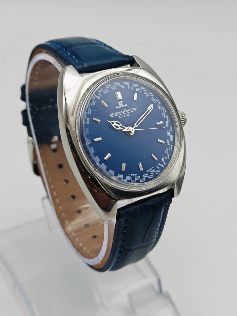 Puede incluir: Un reloj Jaeger-LeCoultre Club vintage con esfera azul y caja plateada. El reloj tiene una correa de cuero azul y una corona plateada. La esfera tiene marcadores de hora y agujas plateadas. El reloj se exhibe sobre un soporte transparente.