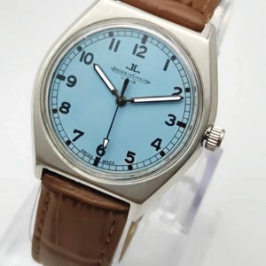 K&ouml;nnte beinhalten: Eine silberfarbene Armbanduhr mit hellblauem Zifferblatt und braunem Lederarmband. Das Zifferblatt zeigt schwarze Ziffern und Zeiger, mit dem Markennamen "JAEGER-LECOULTRE" und "CUB" aufgedruckt. Schweizer Fabrikat.