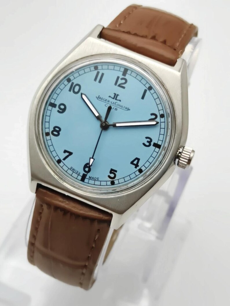 K&ouml;nnte beinhalten: Eine silberfarbene Armbanduhr mit hellblauem Zifferblatt und braunem Lederarmband. Das Zifferblatt zeigt schwarze Ziffern und Zeiger, mit dem Markennamen "JAEGER-LECOULTRE" und "CUB" aufgedruckt. Schweizer Fabrikat.
