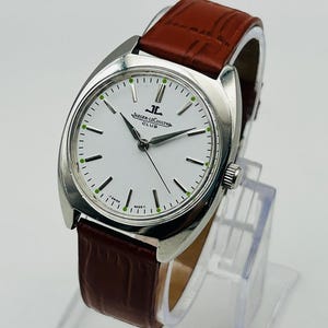 May include: A silver-toned Jaeger-LeCoultre Club watch with a white face and brown leather band. The watch face has black hands and green markers. The band is a rich brown color.