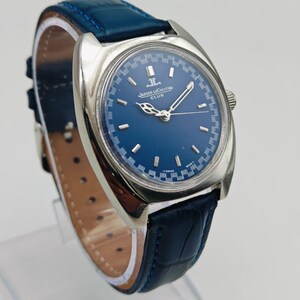 Puede incluir: Un reloj Jaeger-LeCoultre Club plateado con esfera azul y correa de cuero azul. La esfera del reloj tiene marcadores de hora y agujas plateadas. La correa es de cuero azul oscuro texturizado. El reloj se exhibe sobre un soporte transparente.
