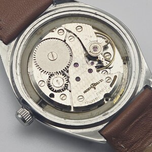 May include: A vintage wristwatch with a silver case and a brown leather strap. The watch's back is open, revealing the intricate mechanical movement with gears, jewels, and the inscription "ULYSSE NARDIN 17 JEWELS".