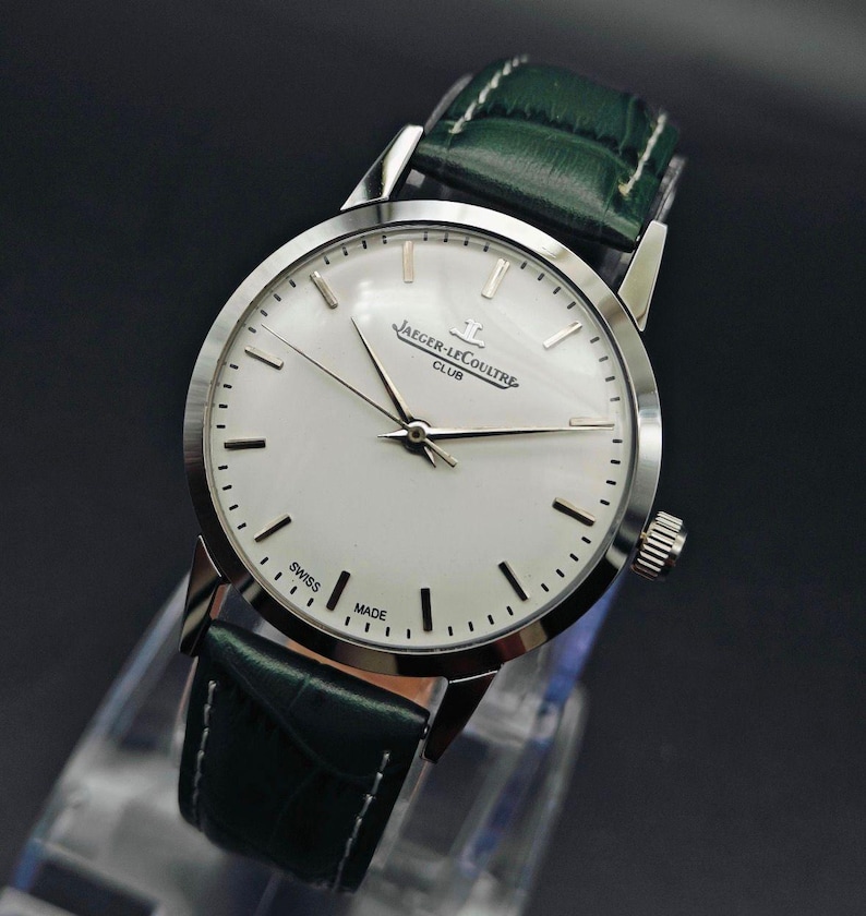 Pu&ograve; includere: Un orologio Jaeger-LeCoultre Club argentato con quadrante bianco e cinturino in pelle verde. L'orologio ha lancette e indici argentati, e le parole "SWISS MADE" sono stampate sul quadrante. L'orologio &egrave; esposto su un supporto trasparente.