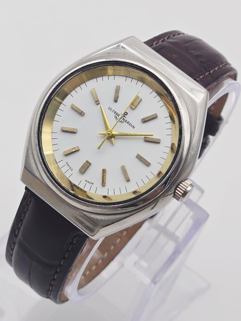 Pu&ograve; includere: Un orologio da polso argentato con quadrante bianco e dettagli dorati. L'orologio presenta un cinturino in pelle marrone e il marchio "ULYSSE NARDIN" &egrave; visibile sul quadrante. Design classico.