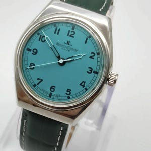 Pode incluir: Um rel&oacute;gio de pulso prateado com mostrador turquesa e pulseira de couro verde escuro. O mostrador exibe o logotipo Jaeger-LeCoultre e n&uacute;meros. A pulseira tem costuras brancas.
