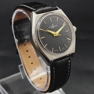 May include: A Ulysse Nardin wristwatch featuring a round black dial with gold-toned hands and hour markers, housed in a silver-toned metal case. The watch is attached to a black leather strap with white stitching and a buckle closure.