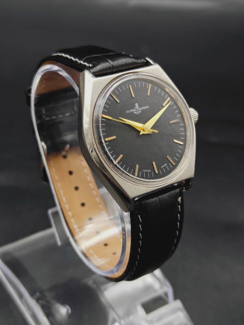 May include: A Ulysse Nardin wristwatch featuring a round black dial with gold-toned hands and hour markers, housed in a silver-toned metal case. The watch is attached to a black leather strap with white stitching and a buckle closure.