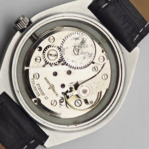 Pu&ograve; includere: Primo piano del complesso meccanismo di un orologio da polso vintage, che mostra ingranaggi, gioielli e componenti meccanici. L'orologio ha una cassa argentata e un cinturino in pelle nera. L'orologio &egrave; marchiato "Ulysse Nardin" e "Seventeen Jewels".