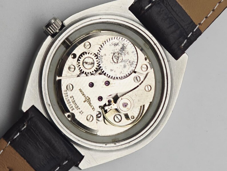 Pu&ograve; includere: Primo piano del complesso meccanismo di un orologio da polso vintage, che mostra ingranaggi, gioielli e componenti meccanici. L'orologio ha una cassa argentata e un cinturino in pelle nera. L'orologio &egrave; marchiato "Ulysse Nardin" e "Seventeen Jewels".