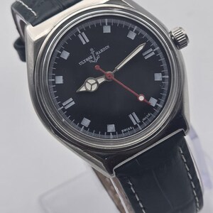 Puede incluir: Un reloj de pulsera Ulysse Nardin de acero inoxidable con esfera negra, marcadores de hora blancos y segundero con punta roja. El reloj tiene una correa de cuero negro con costuras blancas.