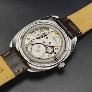 May include: A silver-toned wristwatch with the back casing removed to reveal the internal mechanical movement. The movement is marked Ulysse Nardin 17 Jewels. The watch features a brown leather strap with a light-colored underside.