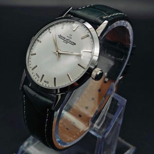 Pu&ograve; includere: Un orologio Jaeger-LeCoultre Club argentato con quadrante chiaro e cinturino in pelle nera. L'orologio ha un quadrante rotondo con lancette e indici argentati. Il cinturino &egrave; verde scuro sulla parte superiore e marrone sul lato inferiore. L'orologio &egrave; esposto su un supporto trasparente.