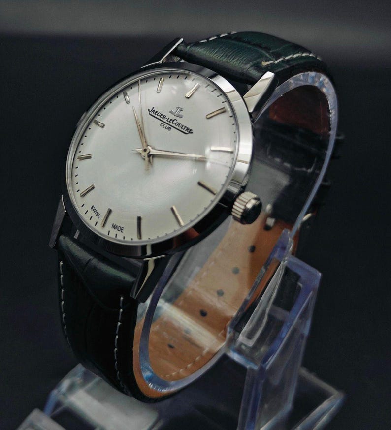 Pu&ograve; includere: Un orologio Jaeger-LeCoultre Club argentato con quadrante chiaro e cinturino in pelle nera. L'orologio ha un quadrante rotondo con lancette e indici argentati. Il cinturino &egrave; verde scuro sulla parte superiore e marrone sul lato inferiore. L'orologio &egrave; esposto su un supporto trasparente.