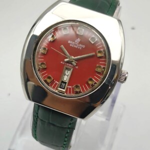 May include: A vintage Breitling Geneve automatic watch with a red dial and silver-tone case. The watch features a date display at the 6 o'clock position and is paired with a green leather strap. The watch has a rectangular shape.