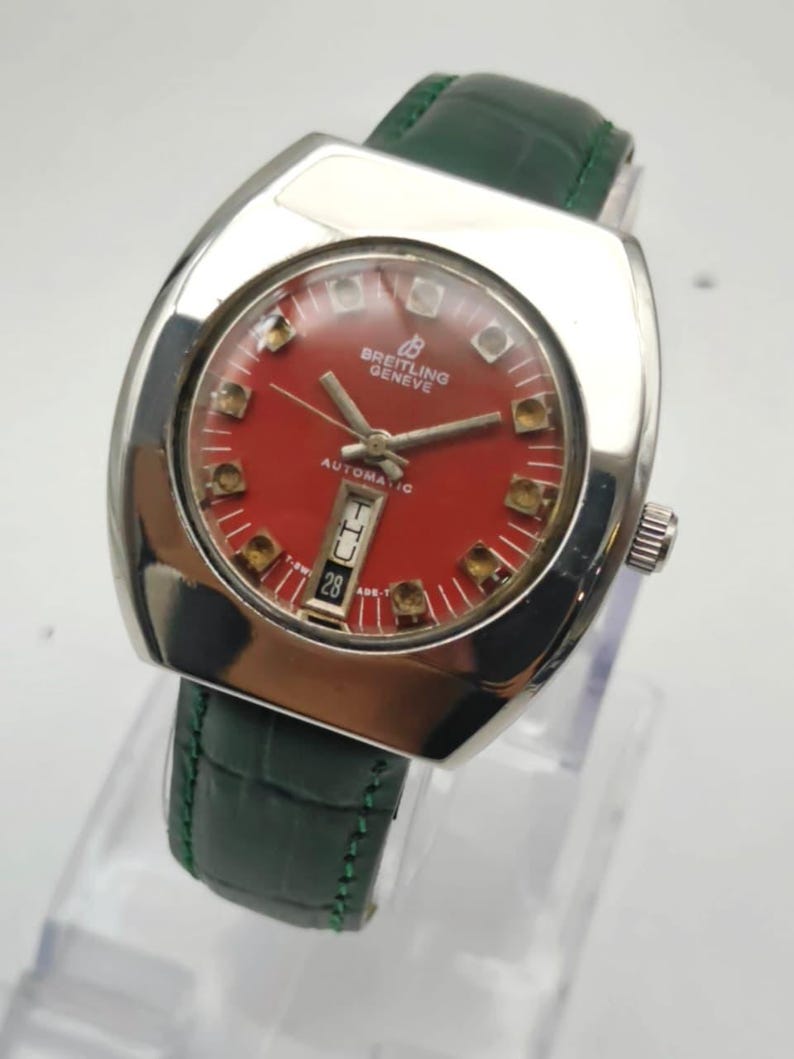 May include: A vintage Breitling Geneve automatic watch with a red dial and silver-tone case. The watch features a date display at the 6 o'clock position and is paired with a green leather strap. The watch has a rectangular shape.