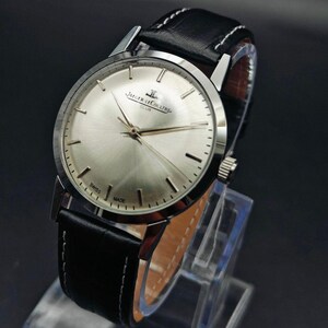 Op de afbeelding: Een zilverkleurig Jaeger-LeCoultre Club horloge met een zwarte leren band en witte stiksels. De wijzerplaat is licht zilverkleurig met goudkleurige wijzers en markeringen. Het horloge wordt op een transparante standaard getoond.