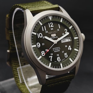 Puede incluir: Un reloj autom&aacute;tico Seiko 5 Sports con caja plateada mate y esfera verde oliva. La esfera presenta n&uacute;meros ar&aacute;bigos blancos, una ventana de d&iacute;a y fecha en la posici&oacute;n de las tres y una correa de tela de nailon verde oliva a juego.