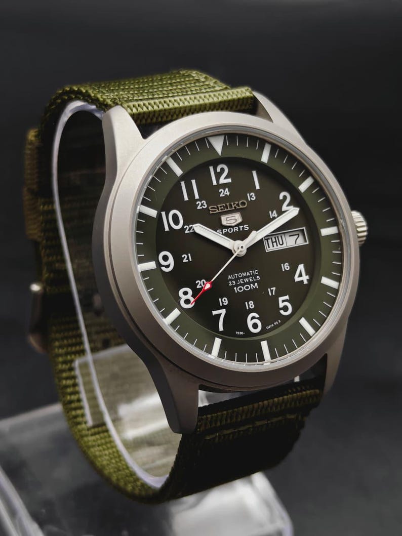 Puede incluir: Un reloj autom&aacute;tico Seiko 5 Sports con caja plateada mate y esfera verde oliva. La esfera presenta n&uacute;meros ar&aacute;bigos blancos, una ventana de d&iacute;a y fecha en la posici&oacute;n de las tres y una correa de tela de nailon verde oliva a juego.
