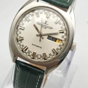 Puede incluir: Un reloj autom&aacute;tico Jaeger-LeCoultre Club plateado con esfera blanca, visualizaci&oacute;n del d&iacute;a y correa de cuero verde. La esfera del reloj presenta el logotipo de Jaeger-LeCoultre y la palabra "AUTOMATIC". El reloj tiene una caja plateada y una correa de cuero verde.